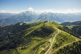 Panoramablick auf den grünen Gipfel der Schmittenhöhe sowie eine schmale Straße, die sich durch die Berglandschaft zieht, mit Bergen im Hintergrund.