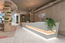 Perspektivischer Blick auf einen modernen Eingangsbereich mit einer holzbekleideten Säule mit Pflanzentrögen links, einem modernen Empfangstresen auf der rechten Seite und Schränken sowie Meeting-Räumen im Hintergrund. © (c) Future Health Lab Perspektivischer Blick auf einen modernen Eingangsbereich mit einer holzbekleideten Säule mit Pflanzentrögen links, einem modernen Empfangstresen auf der rechten Seite und Schränken sowie Meeting-Räumen im Hintergrund.